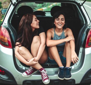 two girls sitting in the back of a car with the boot up and laughing with one another