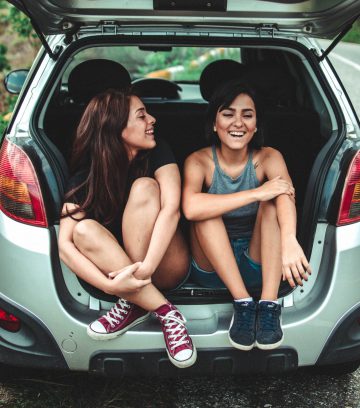 two girls sitting in the back of a car with the boot up and laughing with one another