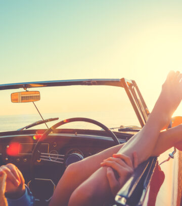 convertible car on a beach with a woman hanging her feet out of the window