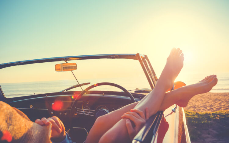 convertible car on a beach with a woman hanging her feet out of the window
