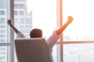happy businessman in office raising fists looking forward to city building urban scene through glass window