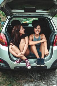 two girls sitting in the back of a car with the boot up and laughing with one another
