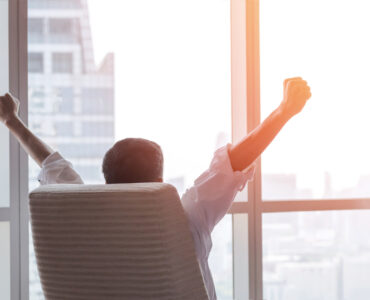 happy businessman in office raising fists looking forward to city building urban scene through glass window