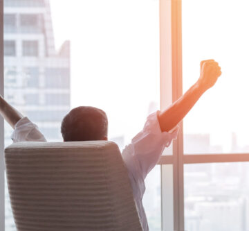 happy businessman in office raising fists looking forward to city building urban scene through glass window