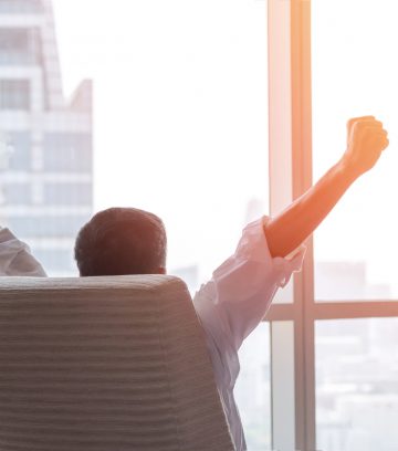 happy businessman in office raising fists looking forward to city building urban scene through glass window