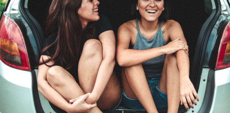 two girls sitting in the back of a car with the boot up and laughing with one another