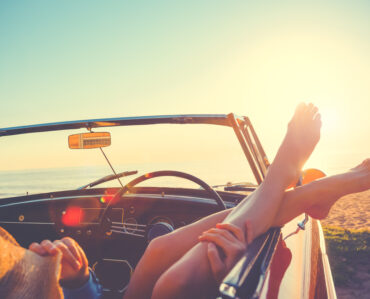 convertible car on a beach with a woman hanging her feet out of the window